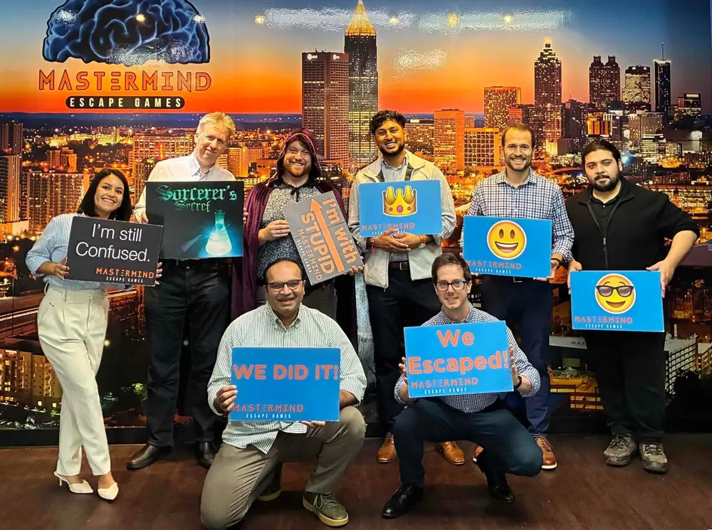 A work team in front of the Atlanta skyline mural holding a Sorcerer’s Secret sign and “We did it!” and “We escaped!” signs, one player in a wizard’s cloak