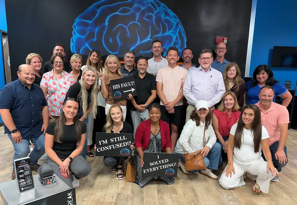 A large corporate group under the Mastermind brain mural, holding “His fault!”, “I’m still confused!?” and “I solved everything!” signs